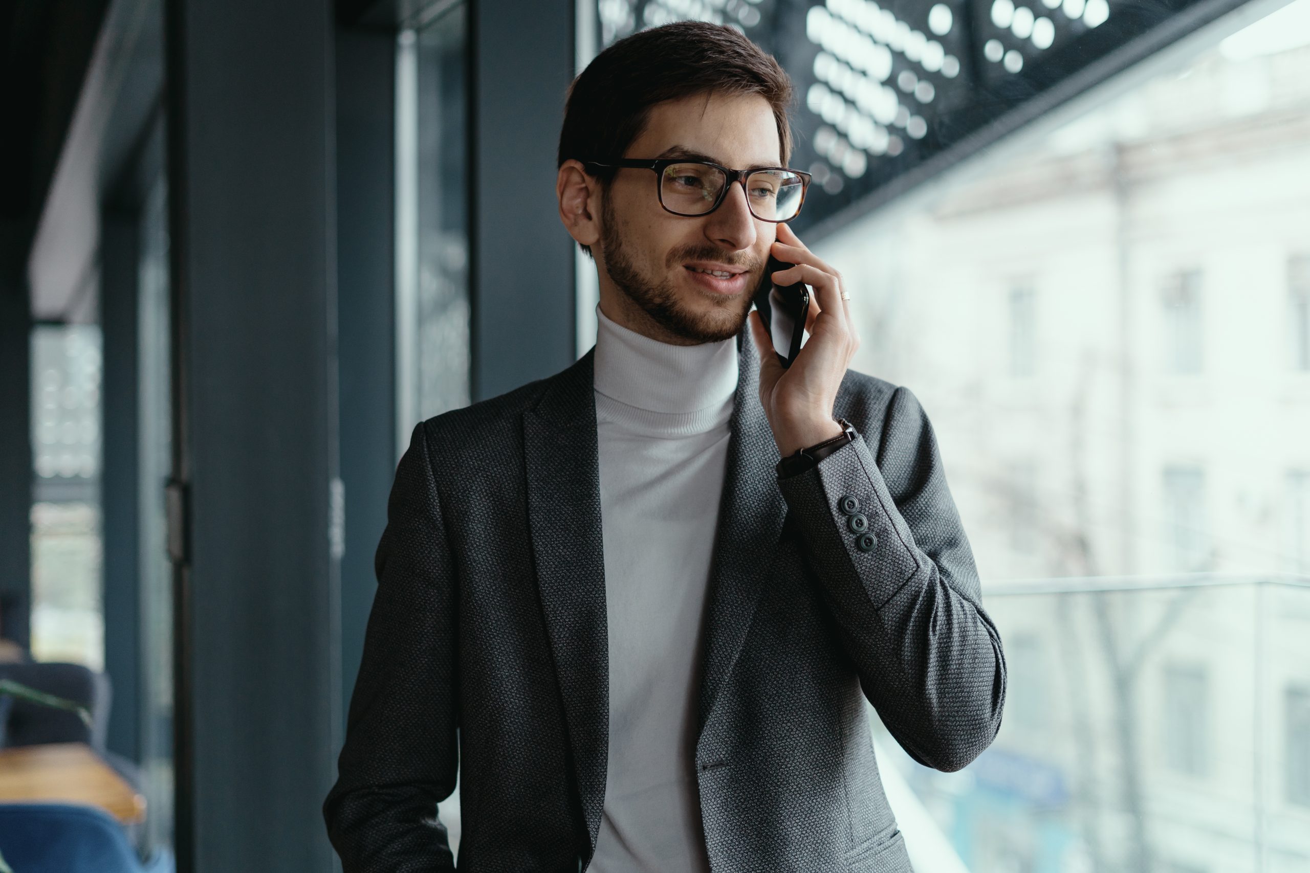 A charismatic young business man wearing glasses, gray jacket, white turtleneck having a conversation on the phone in a modern space